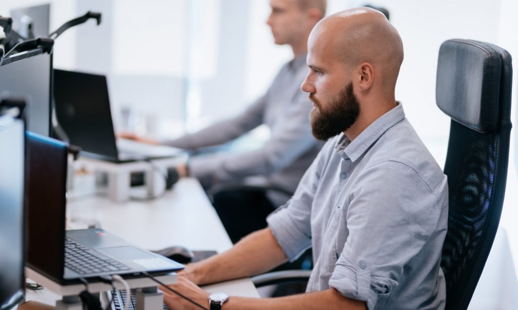 A man with a beard and shaved head sits at a desk working on a computer in an office. Another person, also at a computer, is blurred in the background. Both appear focused on their work.